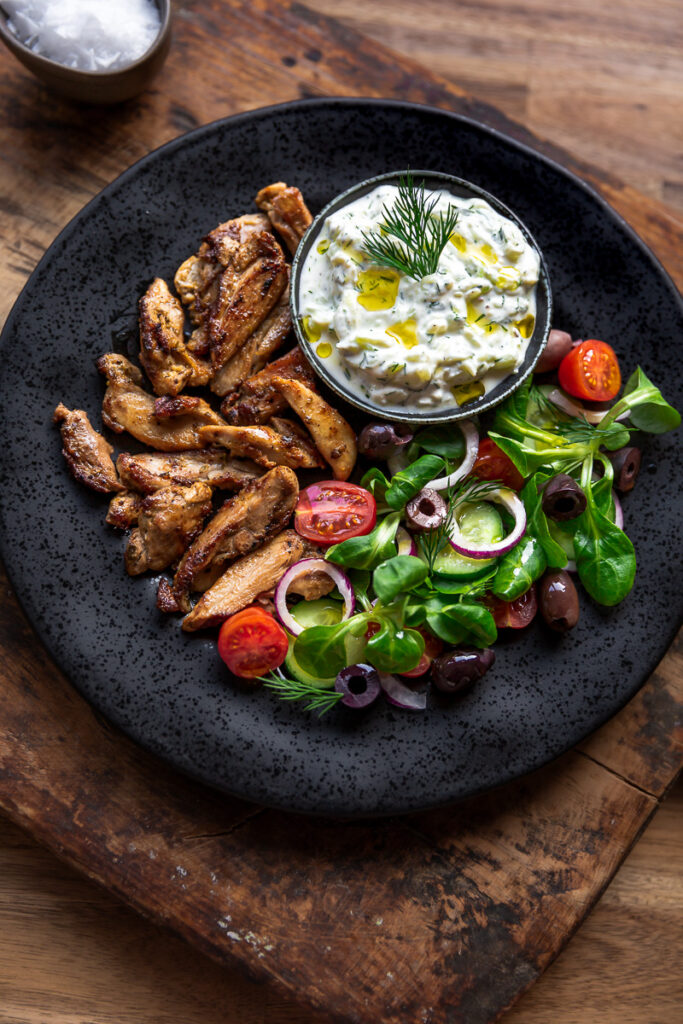 gyros de pui la gratar cu salata si sos tzatziki pe o farfurie neagra pe o masa rustica