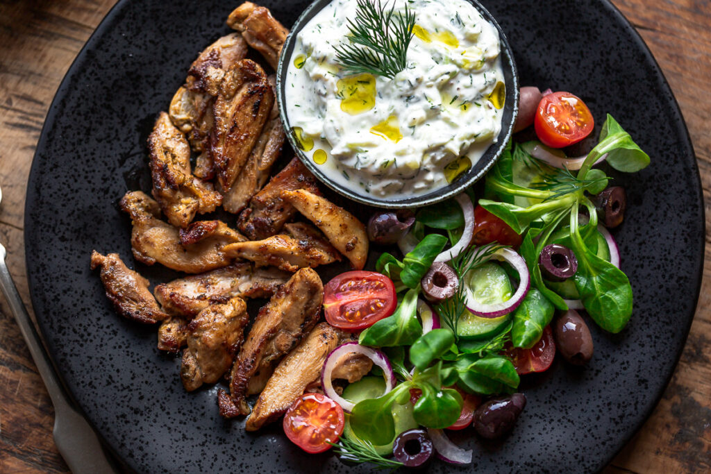 gyros de pui la gratar cu salata si sos tzatziki pe o farfurie neagra pe o masa rustica