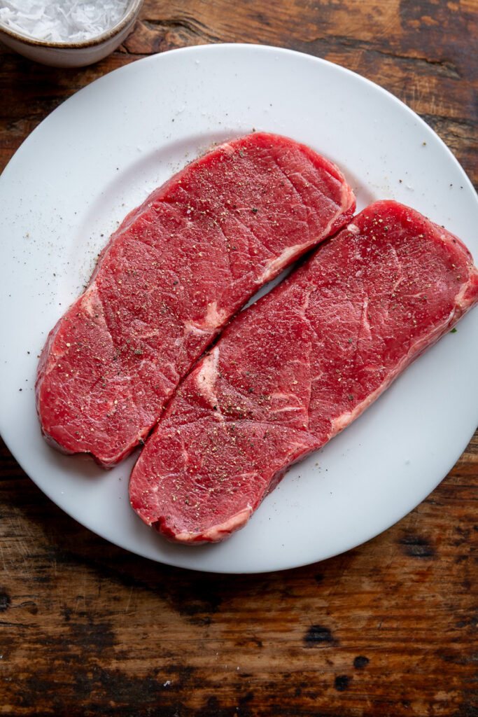 doua felii de carne de vita pe o farfurie alba, cu sare si piper gata de a fi gatite